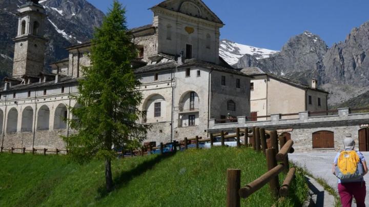 Particolare di fotografia da www.visitcuneese.it, archivio ATL Cuneese Esterno di santuario di San Magno a Castelmagno con loggiati, sfondo di montagne con neve, in visitatore a destra con zainetto in spalla