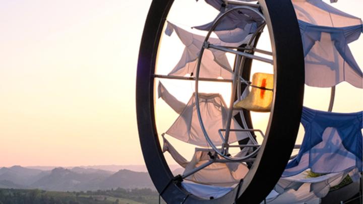 Foto di opera d'arta contemporanea composta da ruota in metallo con elementi di stoffa, sfondo di colline al tramonto