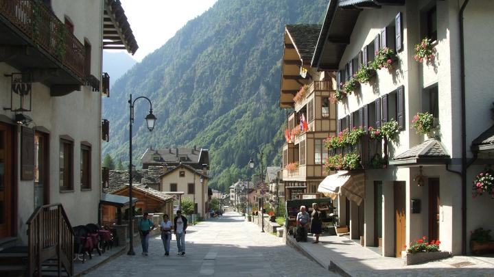 Veduta di Alagna Valsesia, foto da Wikipedia, autori Twice25 & Rinina25 Strada di cittadina in montagna, con sfondo di montagne verdeggianti