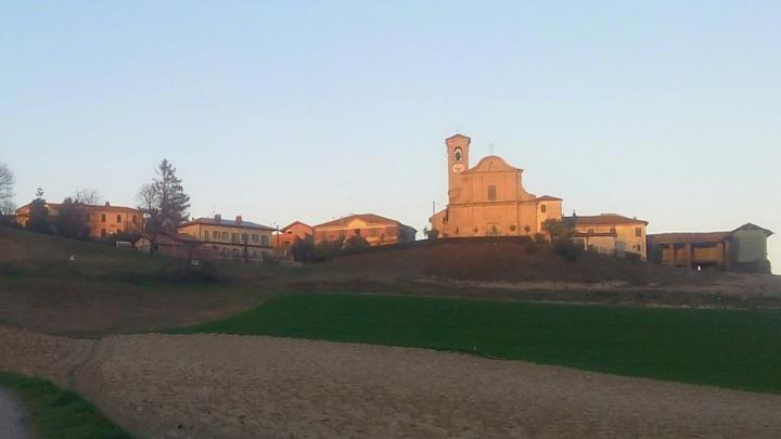 Veduta di borgo collinare con campi in primo piano e chiesa barocca con case sullo sfondo