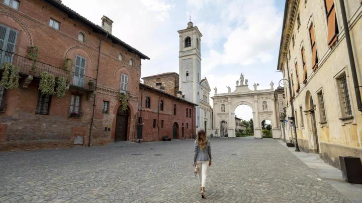Fotografia: "Cherasco_Can't Forget Italy" Archivio Ente Turismo LMR Fotografia con donna che cammina in via di centro storico con arco barocco e campanile sullo sfondo, palazzo gotico a sinistra