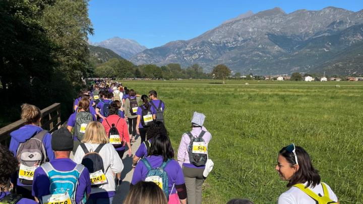 Persone partecipano alla Francigena Marathon camminando tra i prati verdi e le montagne sullo sfondo che si stagliano su un cielo limpido