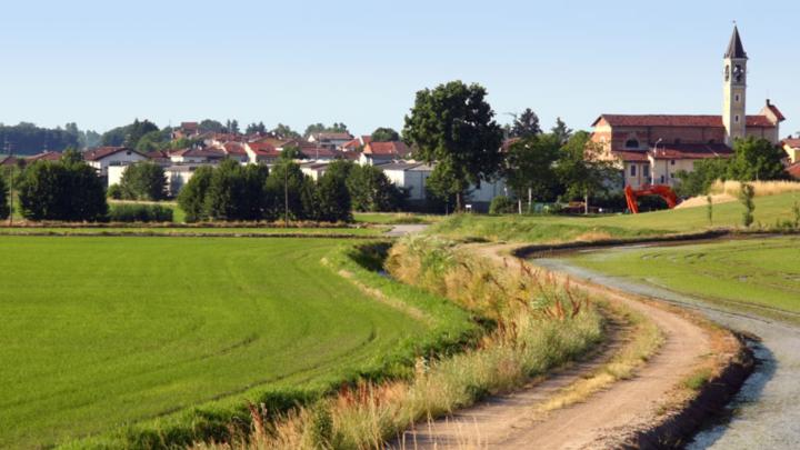 veduta fotogrfica con piccolo comune con case, chiesa e campanile vista dai prati e campi agricoli circostanti, in primo luogo un sentiero