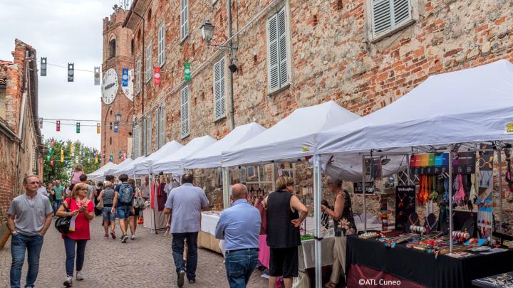 Delle persone passeggiano tra le bancarelle bianche collocate lungo la parete esterna di un palazzo in pietra.