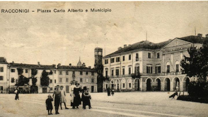 Particolare di antica cartolina di Racconigi d'inizio '900, dal sito /www.cuneoalps.it Foto in bianco e nero con gruppo di adulti e fanciulli in abiti del primo Novecento in una piazza con palazzo neoclassico e alberi