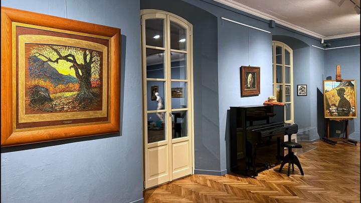 Allestimento della mostra con quadro di paesaggio in primo piano, sullo sfondo pianoforte e altri dipinti