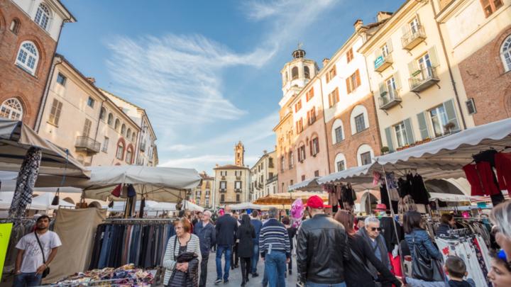 Mercato con bancarelle e folla in una piazza di Savigliano con palazzi e torri medievali in mattoni