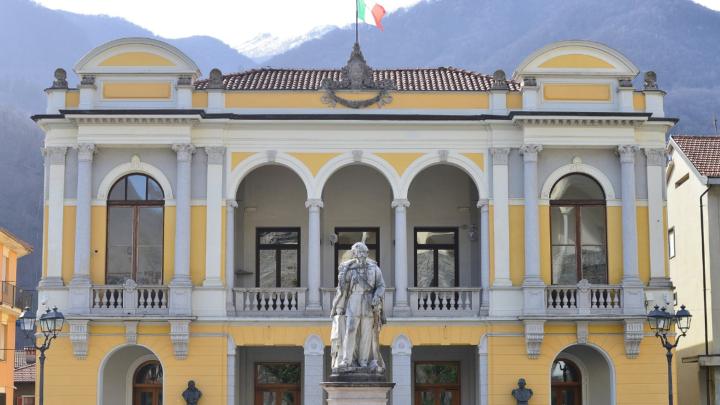 Piazza con facciata di teatro ottocentesco in stile eclettico, in primo piano statua, sullo sfondo le Alpi