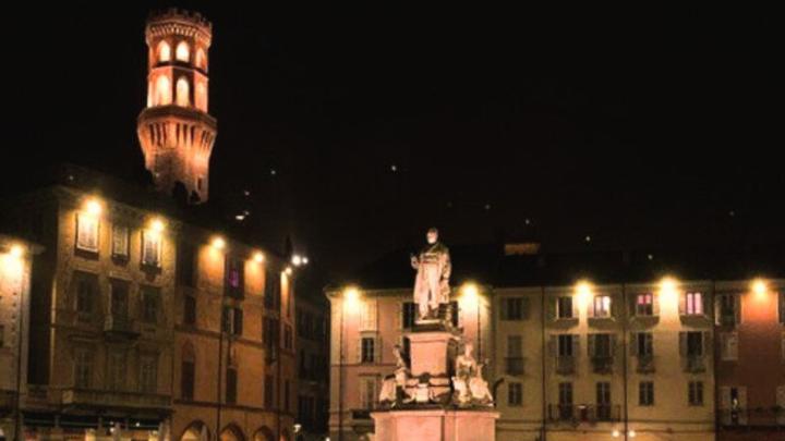 Veduta fotografica di Piazza Cavour a Vercelli di sera, particolare, da www.arteemusei.com Piazza porticata di sera con torre ottagonale e monumento centrale