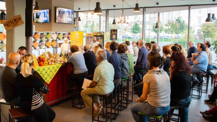 Show cooking all'Open Baladin di Cuneo: prodotti agricoli e gastronomici su un bancone in un ambiente con vetrate, con numeroso pubblico seduto su sgabelli